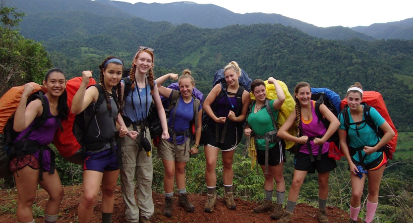 A group of people wearing backpacks flex their arms for the photo. There are tree-covered mountains in the background. 